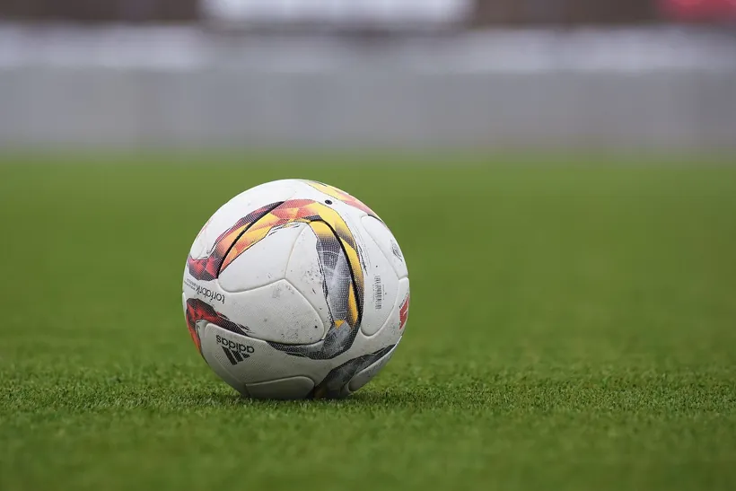 Bola de jogo usada, branca com painéis laranja, pousada na relva com o fundo desfocado