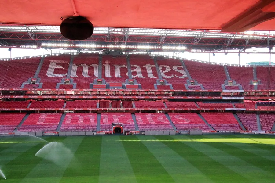 Interior vazio do Estádio da Luz com os aspersores a regar o relvado e as cadeiras vermelhas da bancada em frente