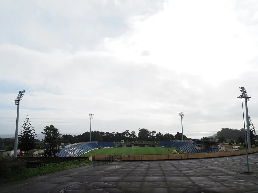Estádio de São Miguel visto de um parque de estacionamento, com bancadas azuis baixas, quatro torres de iluminação e o mar ao fundo